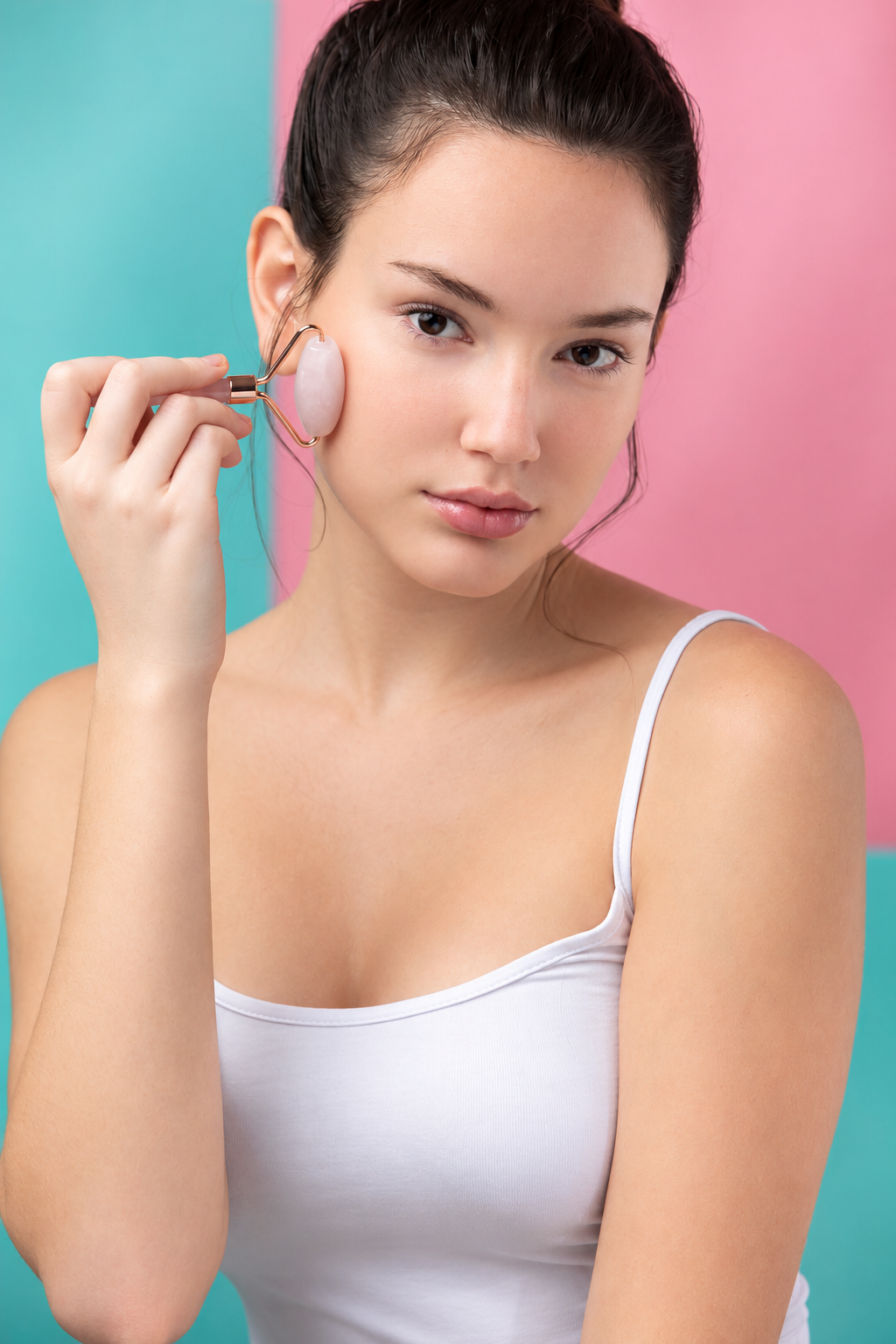 A woman with dark hair tied up is shown from the chest up, using a pink jade facial massage tool on her cheek. She is wearing a white spaghetti-strap tank top against a split pastel teal and pink background.